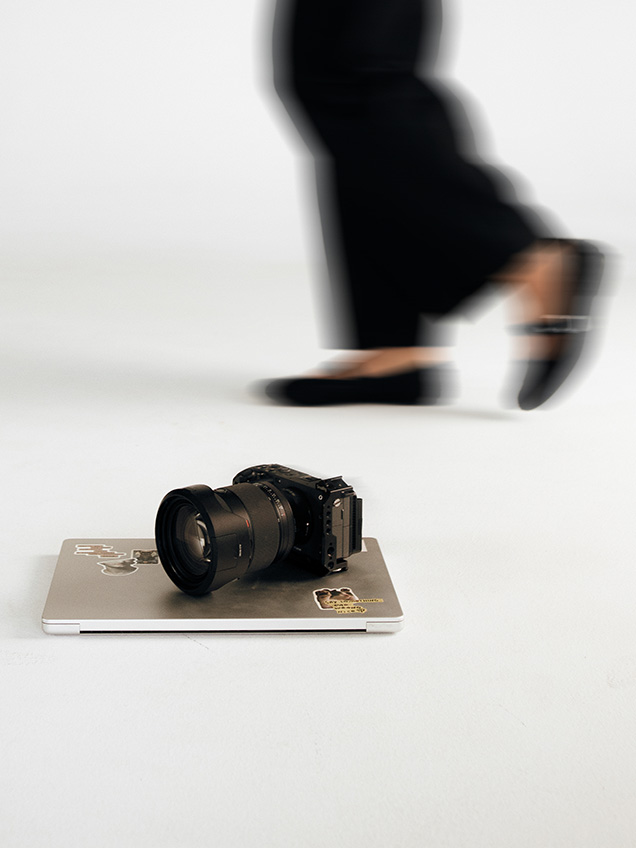 Camera resting on a closed laptop on a white studio floor, with blurred legs walking past in the background.