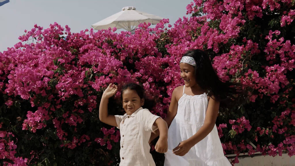 Eddy and Faye’s two children smiling and playing in front of bright pink bougainvillea in Oia.