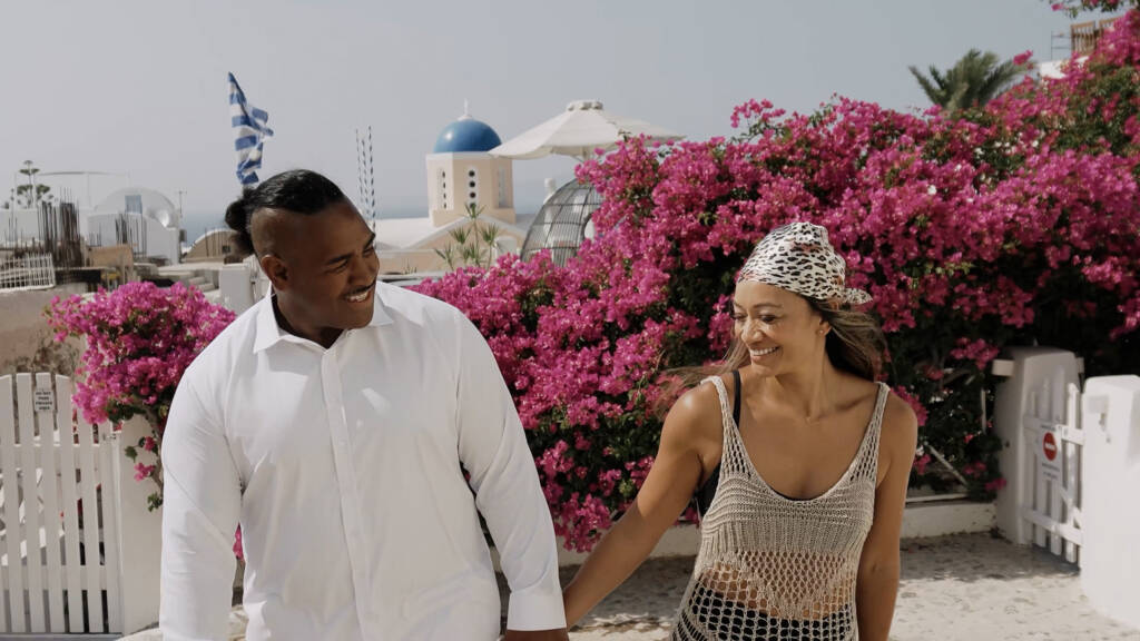 Eddy and Faye smiling at each other as they walk past pink bougainvillea in Oia, Santorini.