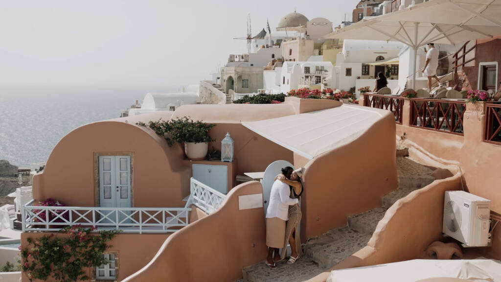 Eddy and Faye embracing on a stone stairway beside peach coloured buildings in Oia, with the sea and windmills behind them.