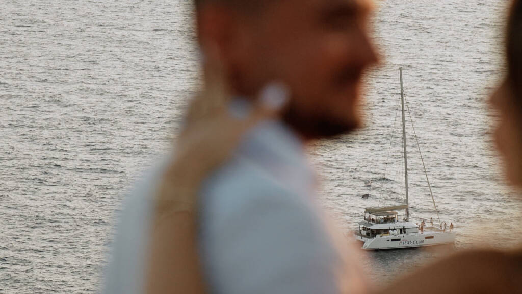 A catamaran sailing off Santorini, with Blendi and Aldona softly out of focus in the foreground.