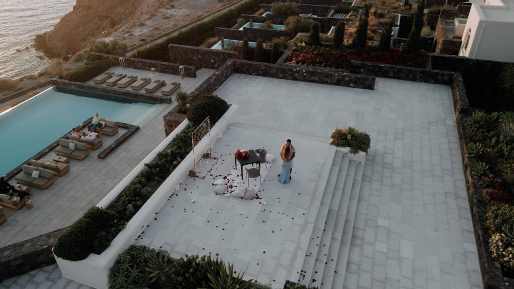 Aerial view of Blendi and Aldona embracing beside a dinner table and scattered rose petals on a Canaves Epitome terrace.