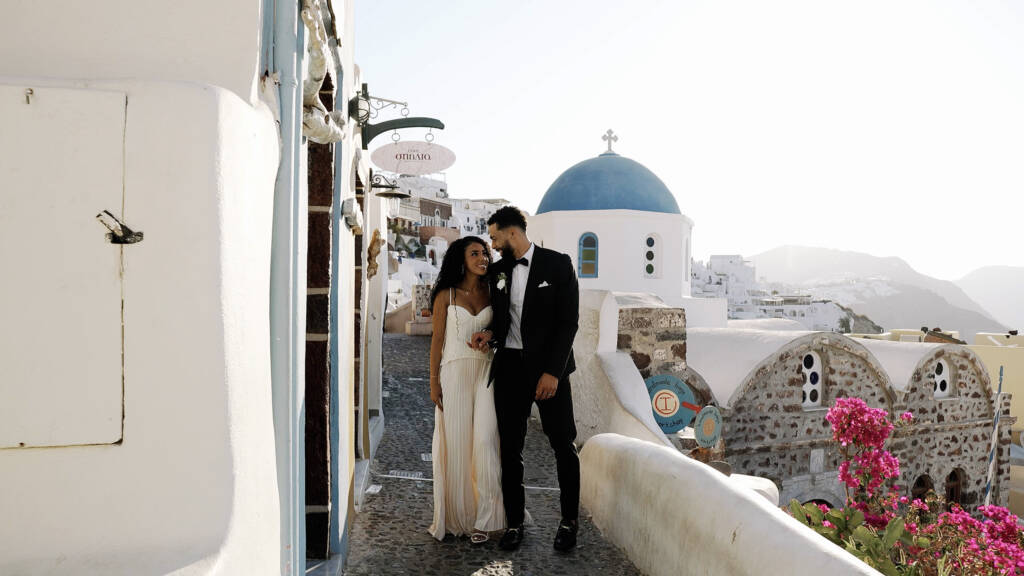 Beeminet looking at Donovan during their sunrise vow exchange, with Oia’s whitewashed buildings behind her.