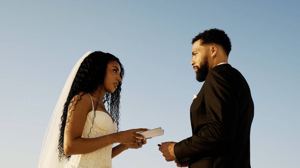 Beeminet looking at Donovan during their sunrise vow exchange, with Oia’s whitewashed buildings behind her.